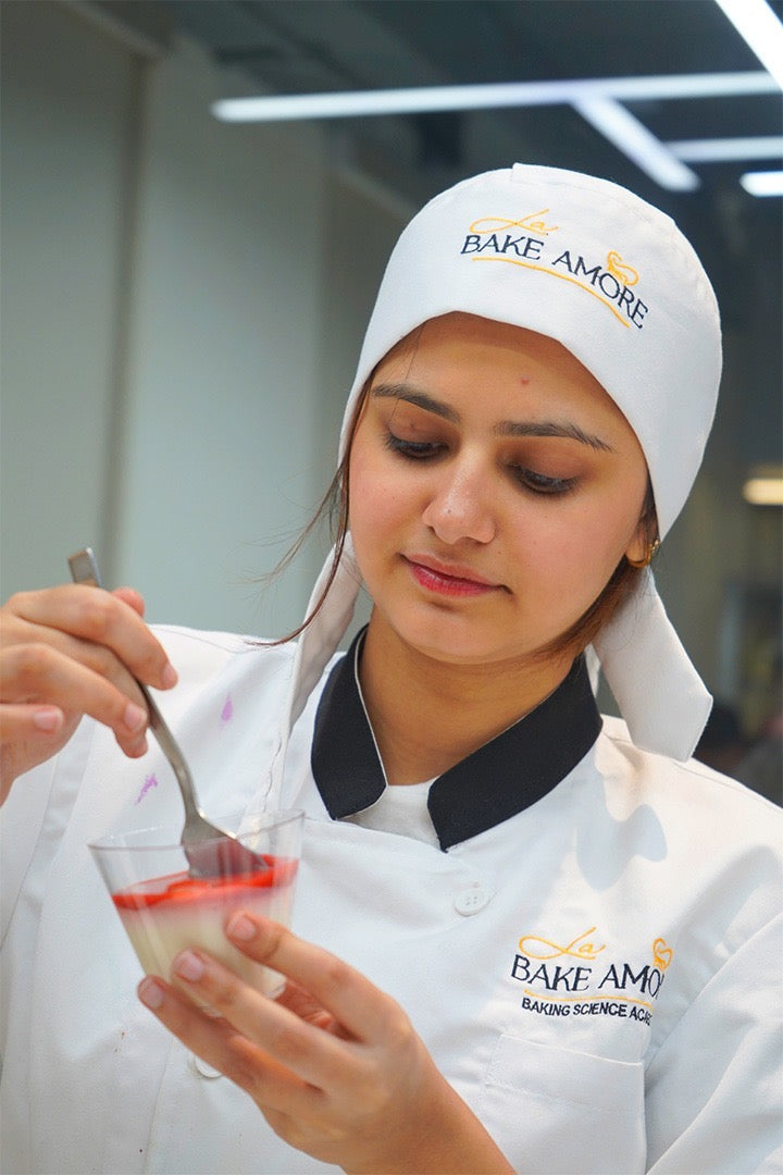 Person in a kitchen wearing a 'Bake Amore' chef's hat and apron, holding a small dish with a spoon at 'La Bake Amore' Delhi.