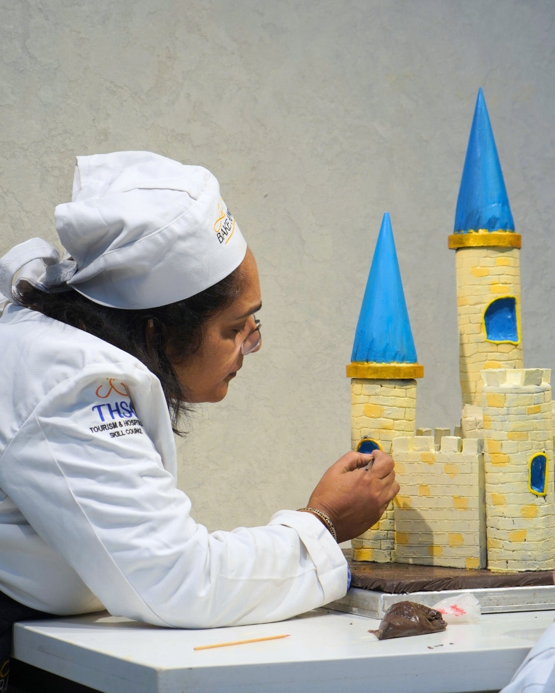 Person in a chef's uniform working on a cake decorated like a castle with blue and yellow towers.