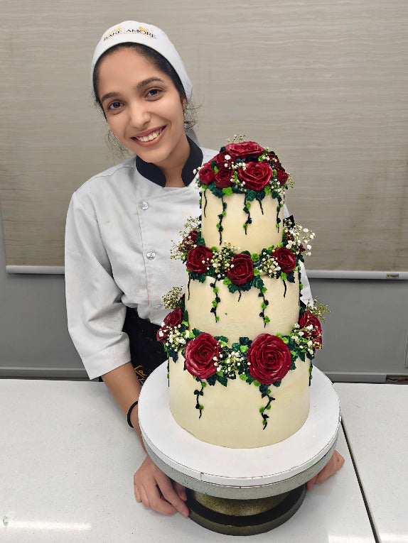Person in a chef's outfit with a three-tiered cake decorated with red roses and greenery at 'La Bake Amore' Delhi.