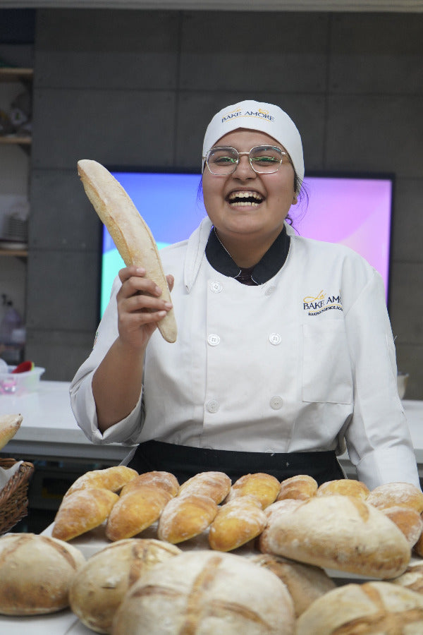 Person in a chef's uniform holding a loaf of bread with bread displayed on shelves in the background.