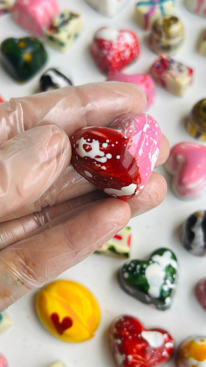 Hand holding a colorful heart-shaped chocolate with more chocolates in the background.