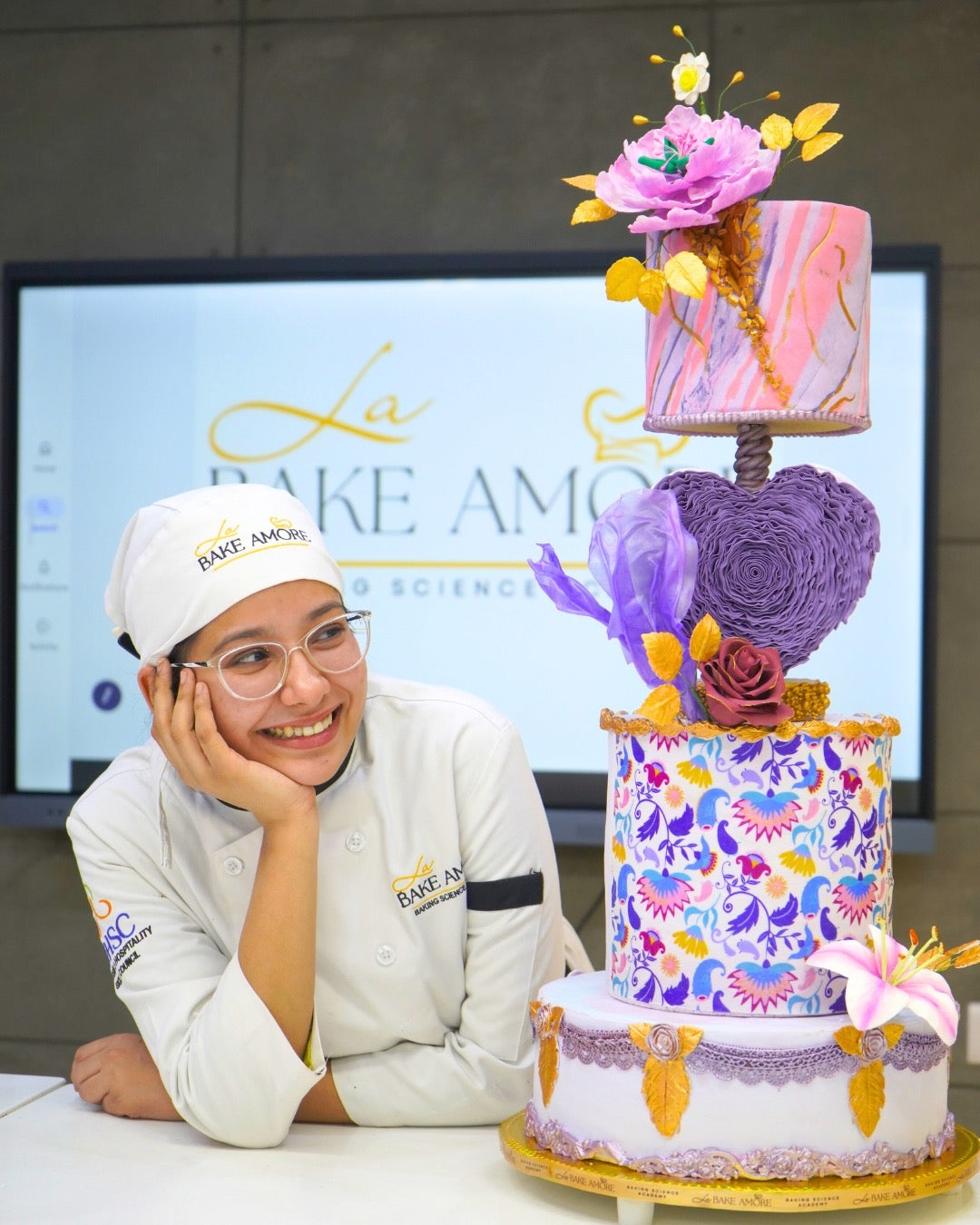 Student in a chef's uniform next to a colorful cake with decorative elements
