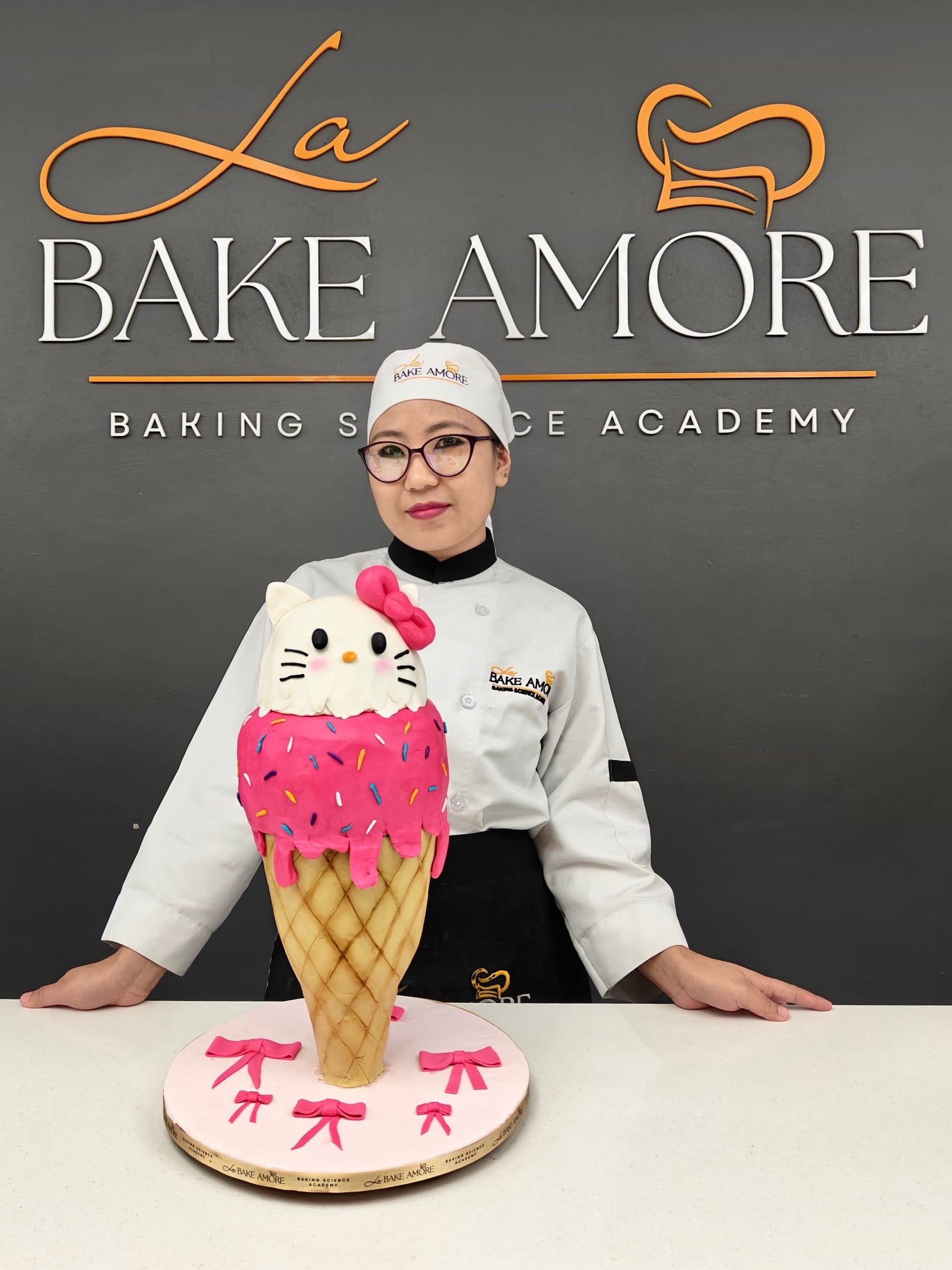 Person in a chef's uniform standing behind a cake shaped like a ice cream cone with a character face, branded 'La Bake Amore'.