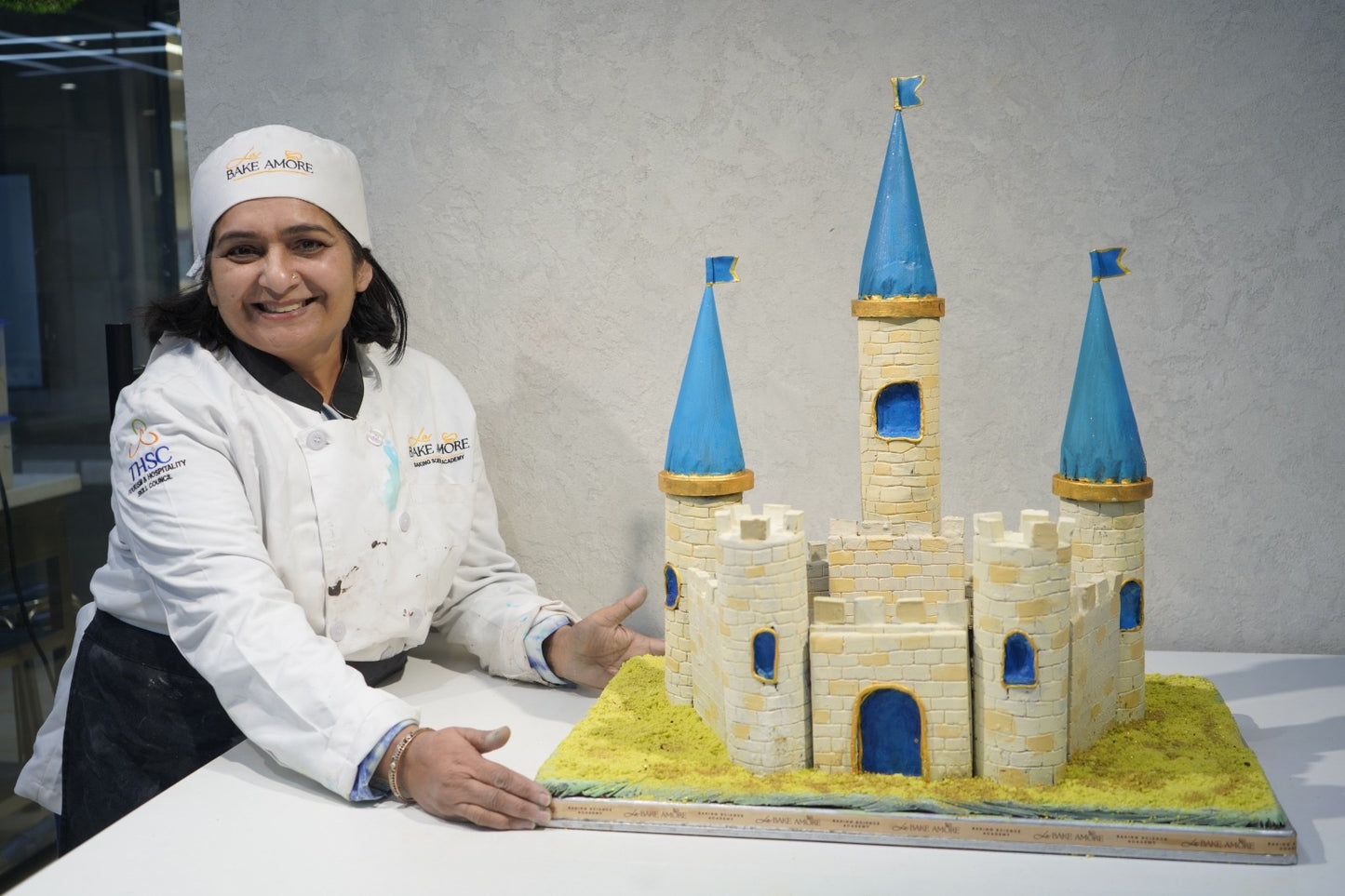 Person in a chef's uniform next to a cake designed like a castle with blue towers.