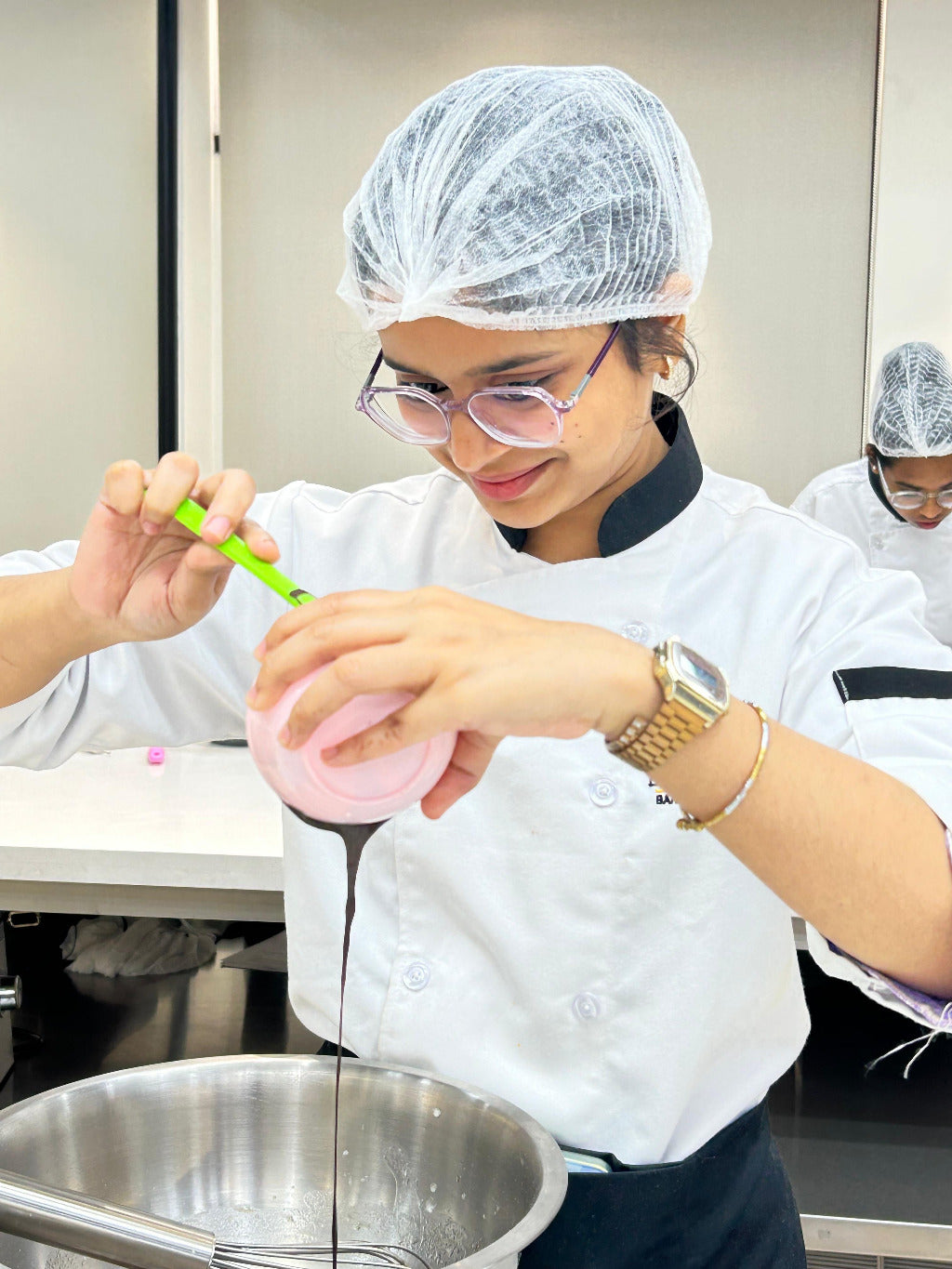 Person in a kitchen wearing a hairnet and chef's coat, pouring liquid from a pink container into a pot.