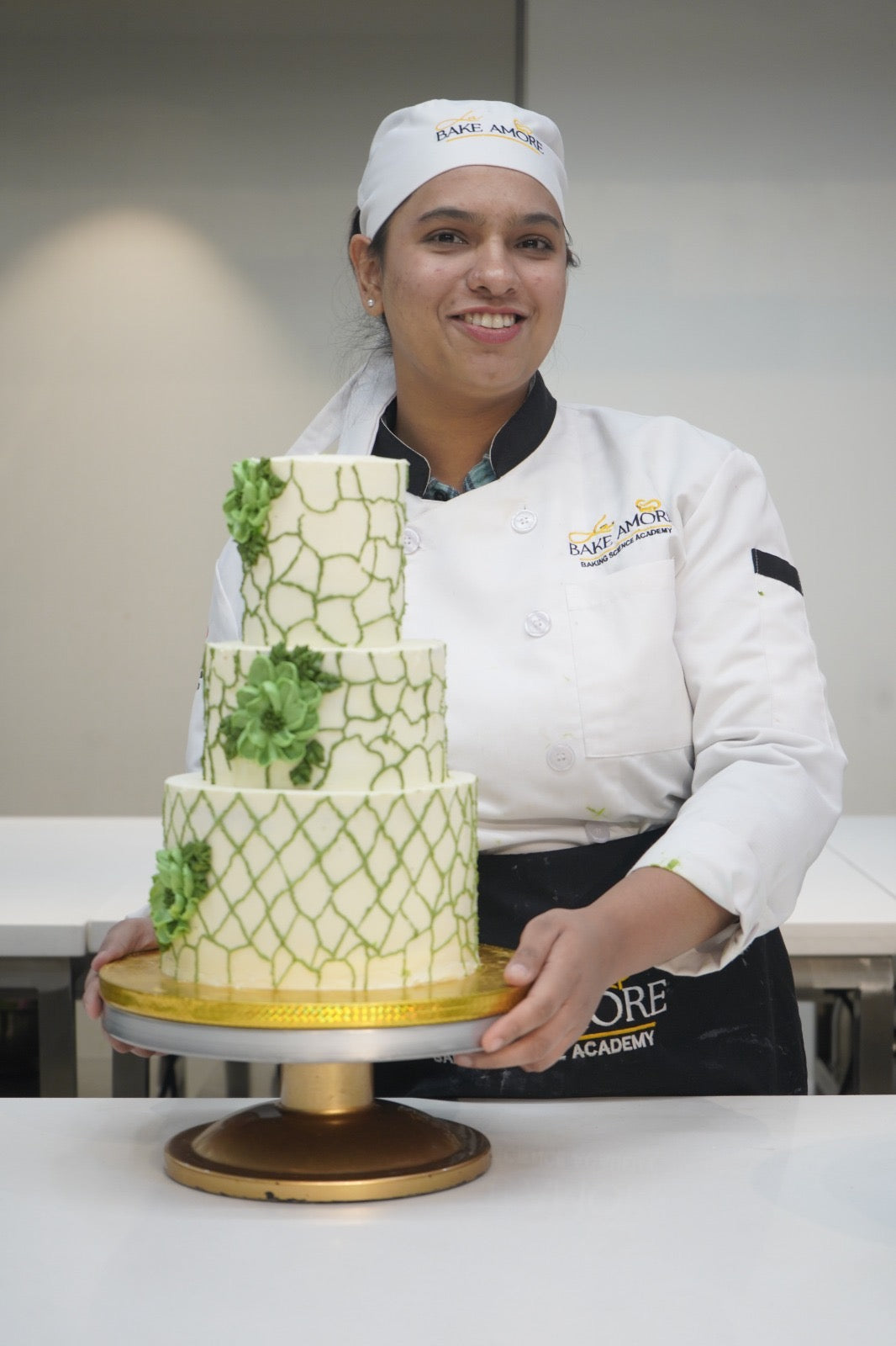 Person in a chef's uniform holding a four-tiered cake with green patterns