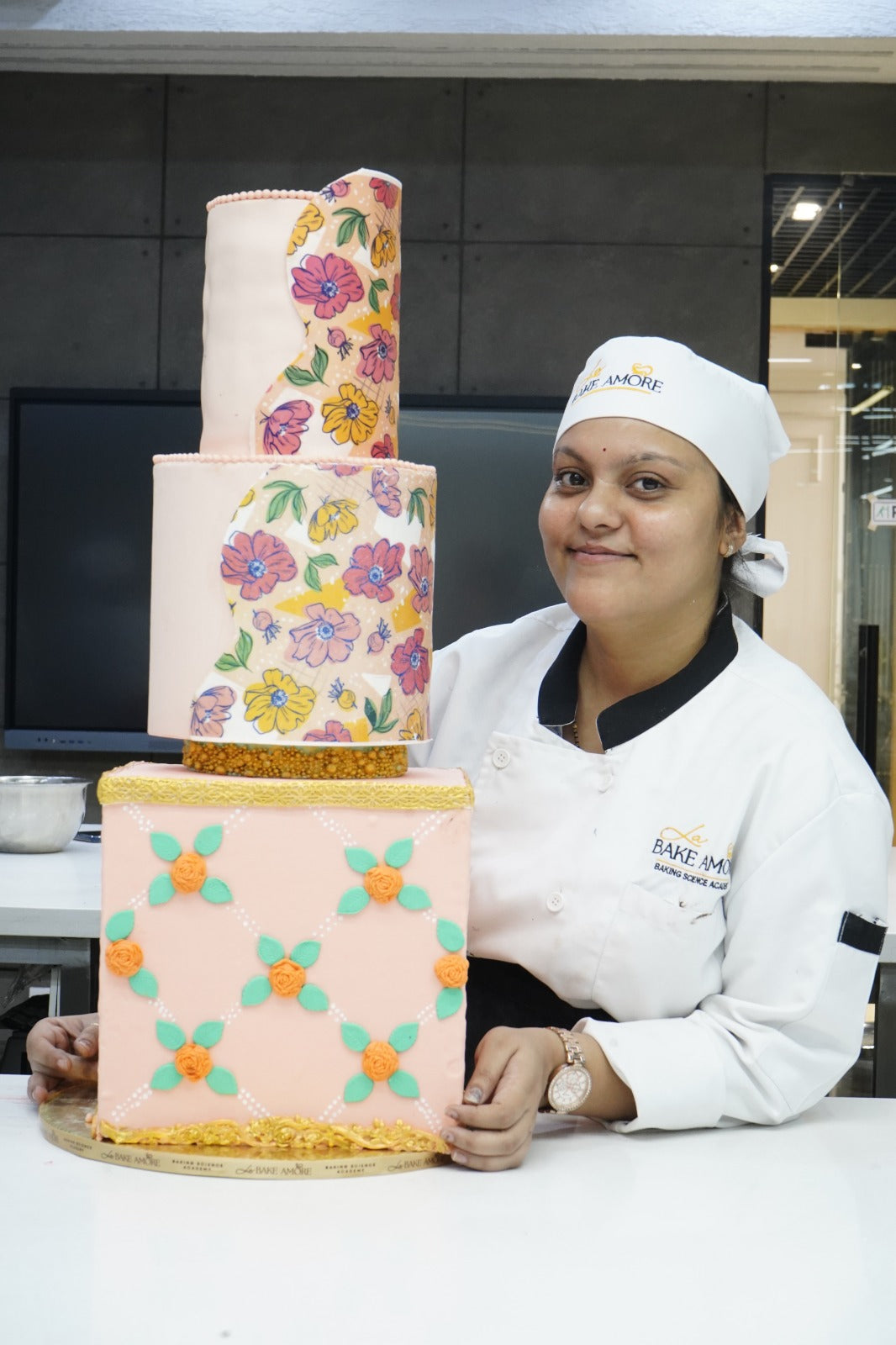 Baker posing with a three-tiered cake decorated with floral patterns in a kitchen setting.